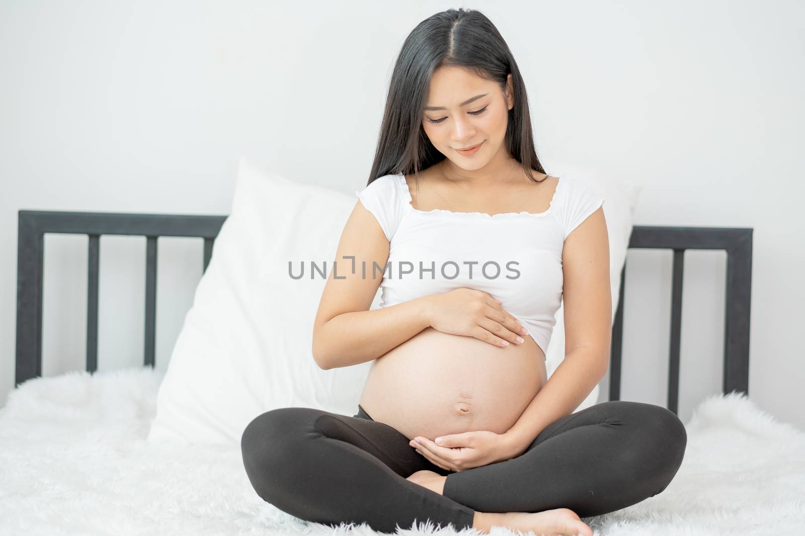 Portrait of beautiful Asian pregnant woman sit on bed and touch and look to her belly also smiling. Concept of good take care baby in the womb of healthy mother and stay at home.