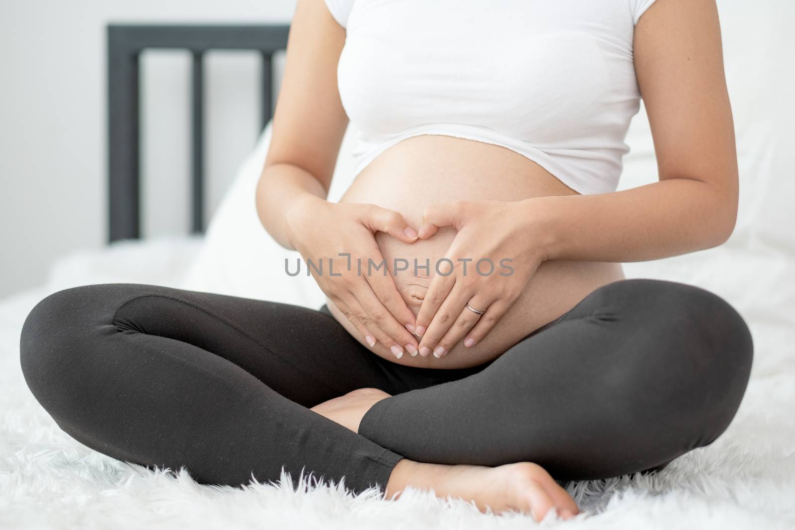 Close up pregnant woman hands touch her belly and make symbol of heart also sit on white bed with morning light. Concept of good take care baby in the womb of healthy mother and stay at home.