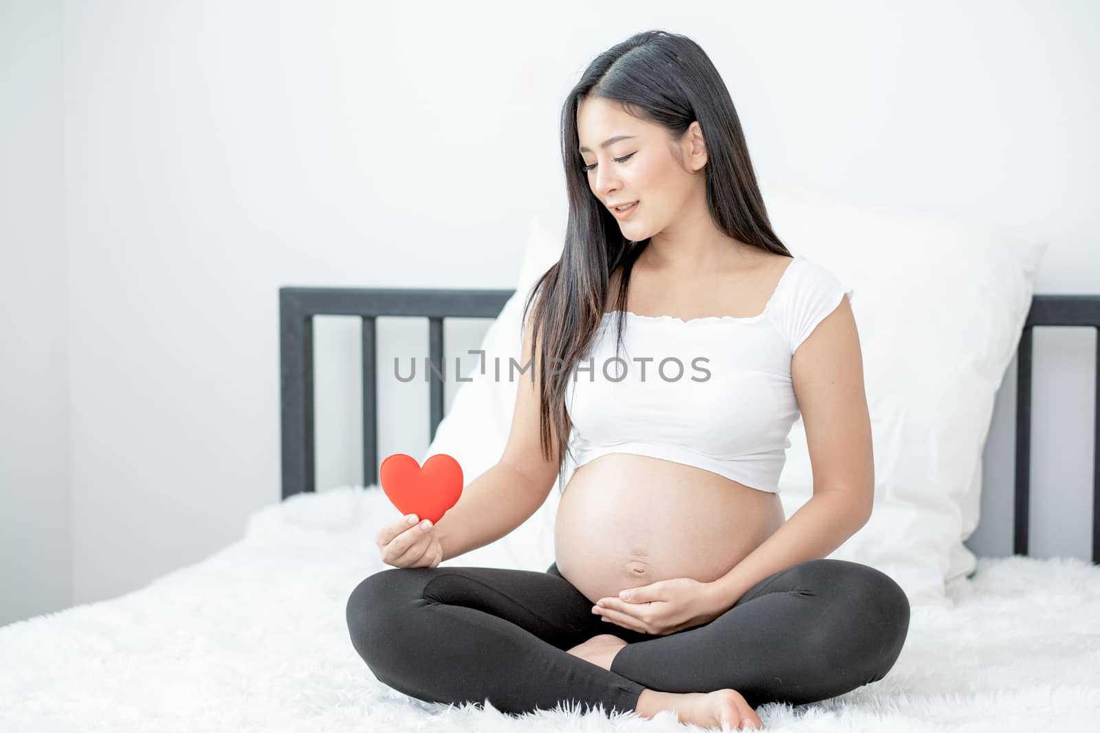 Portrait of beautiful Asian pregnant woman sit on bed and hold red heart icon also smiling and look to symbol. Concept of good take care baby in the womb of healthy mother and stay at home.