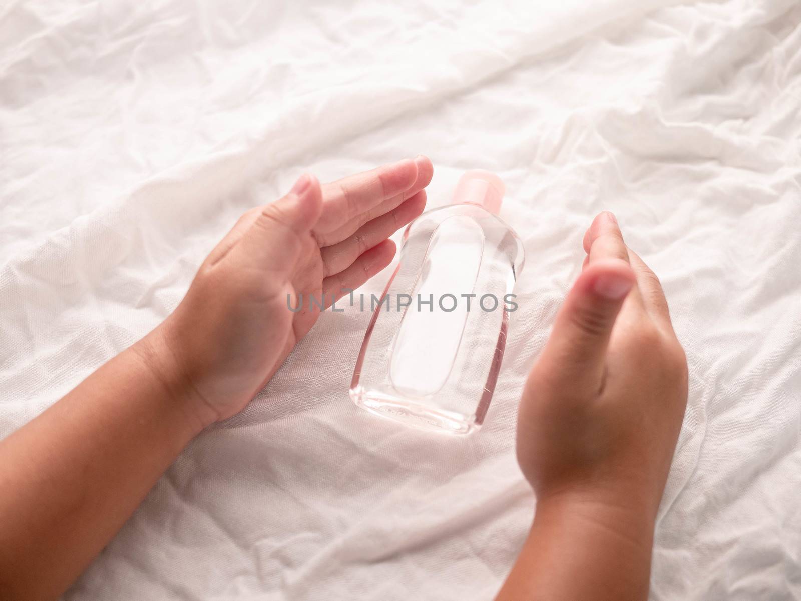 Close-up of Little hands holding transparent plastic bottle of oil on white fabric background. Care about soft body baby skin.