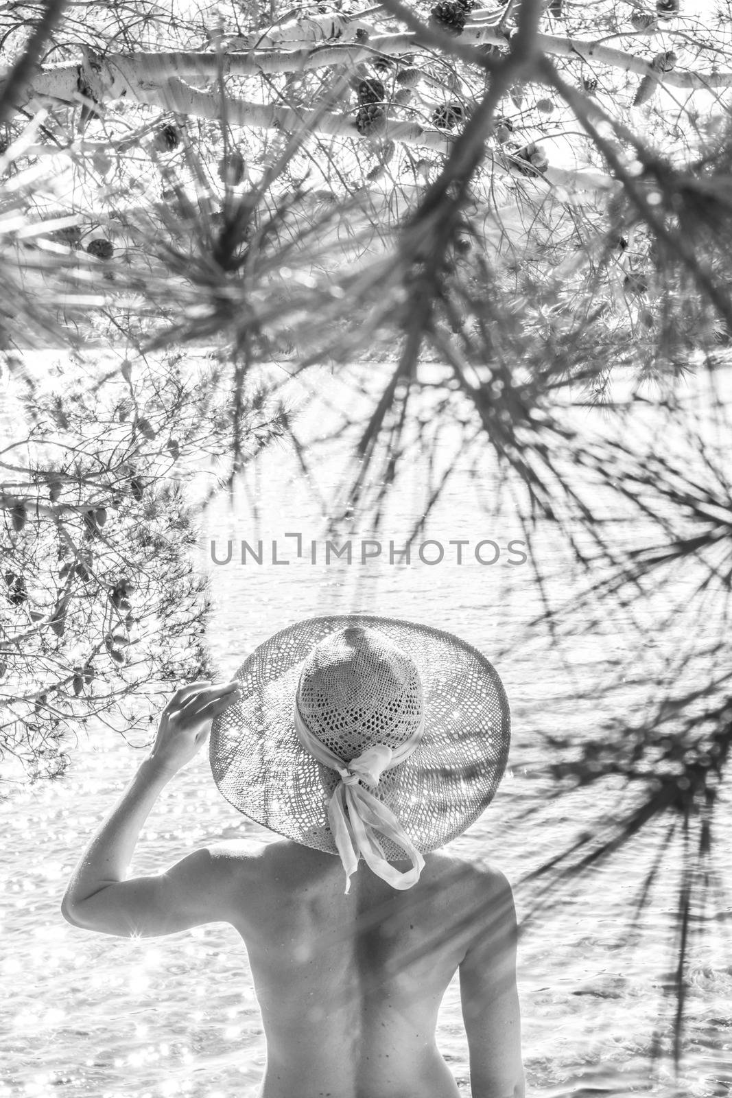 Rear view of topless beautiful woman wearing nothing but straw sun hat realaxing on wild coast of Adriatic sea on beach in shade of pine tree. Relaxed healthy lifestyle concept. Black and white image by kasto
