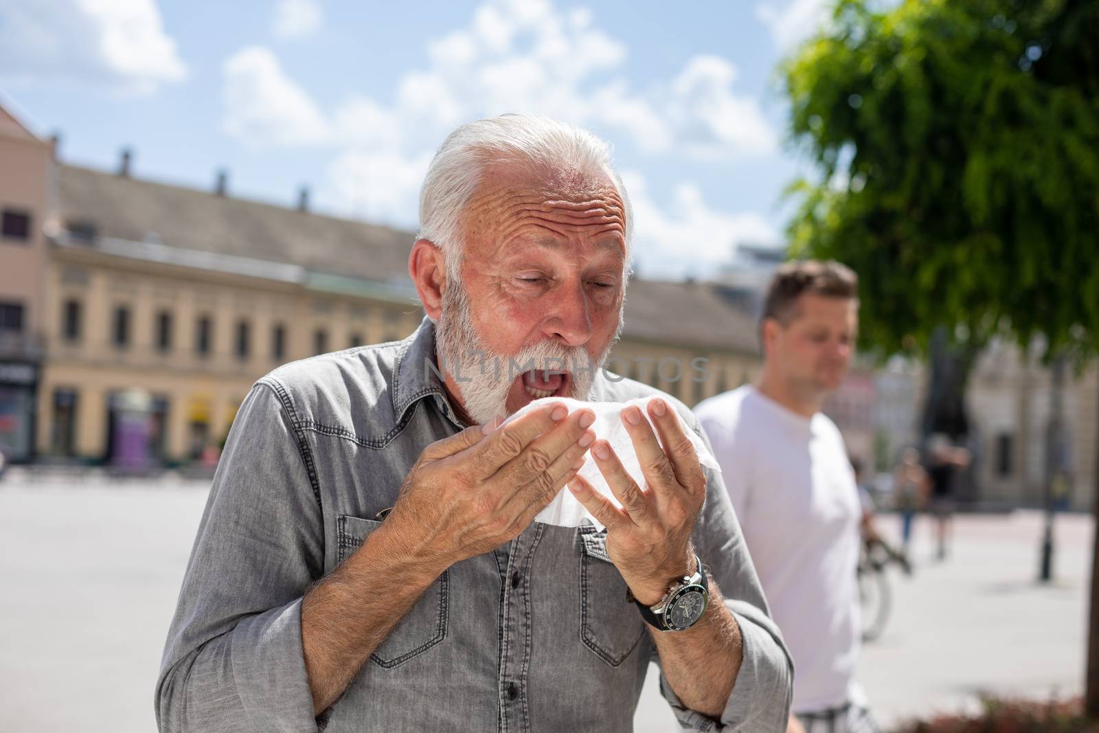 Old man man coughs and sneezes into a handkerchief on street, al by adamr