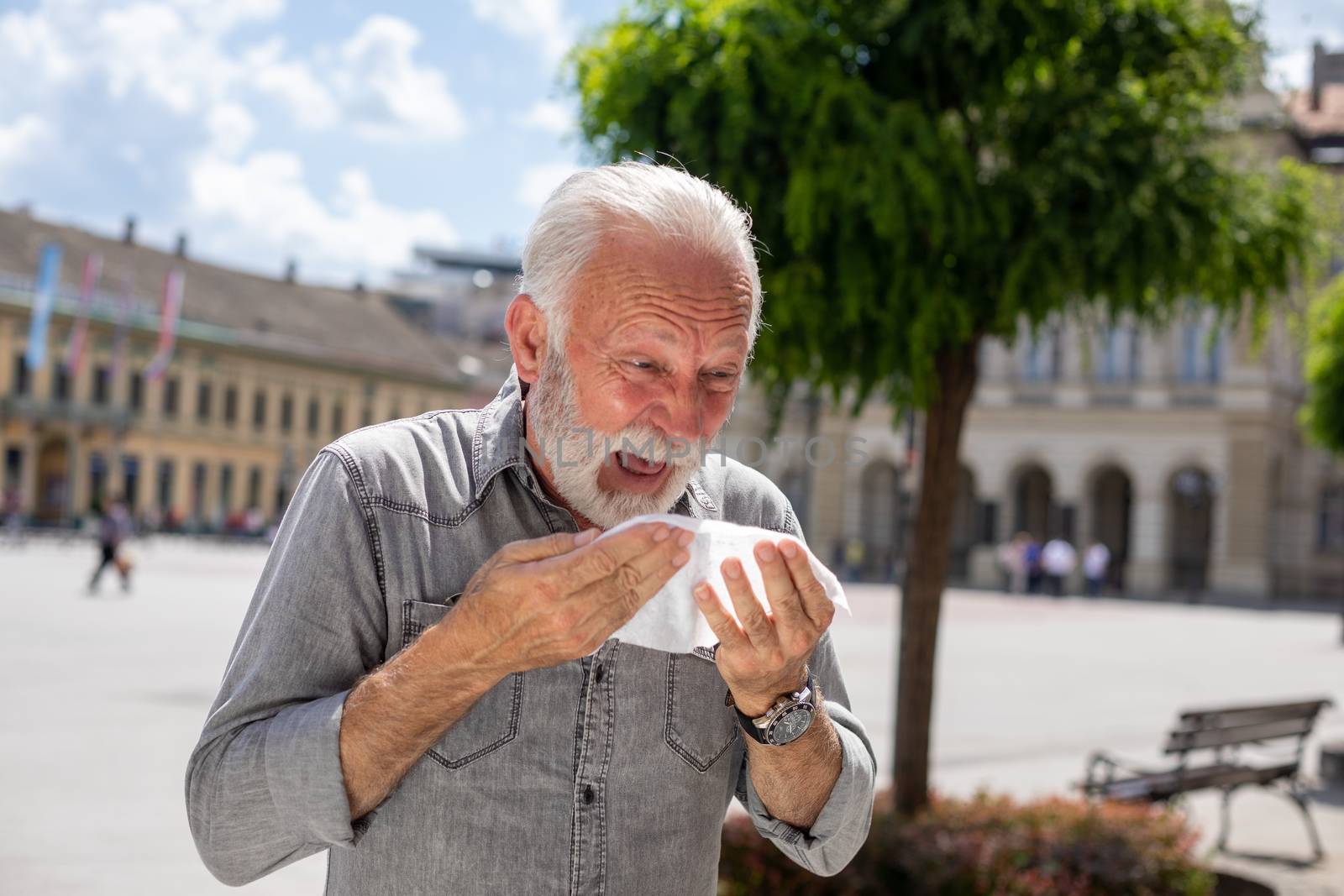 Old man coughs and sneezes into a handkerchief on street, outdoor, hot summer, allergies and illness concept