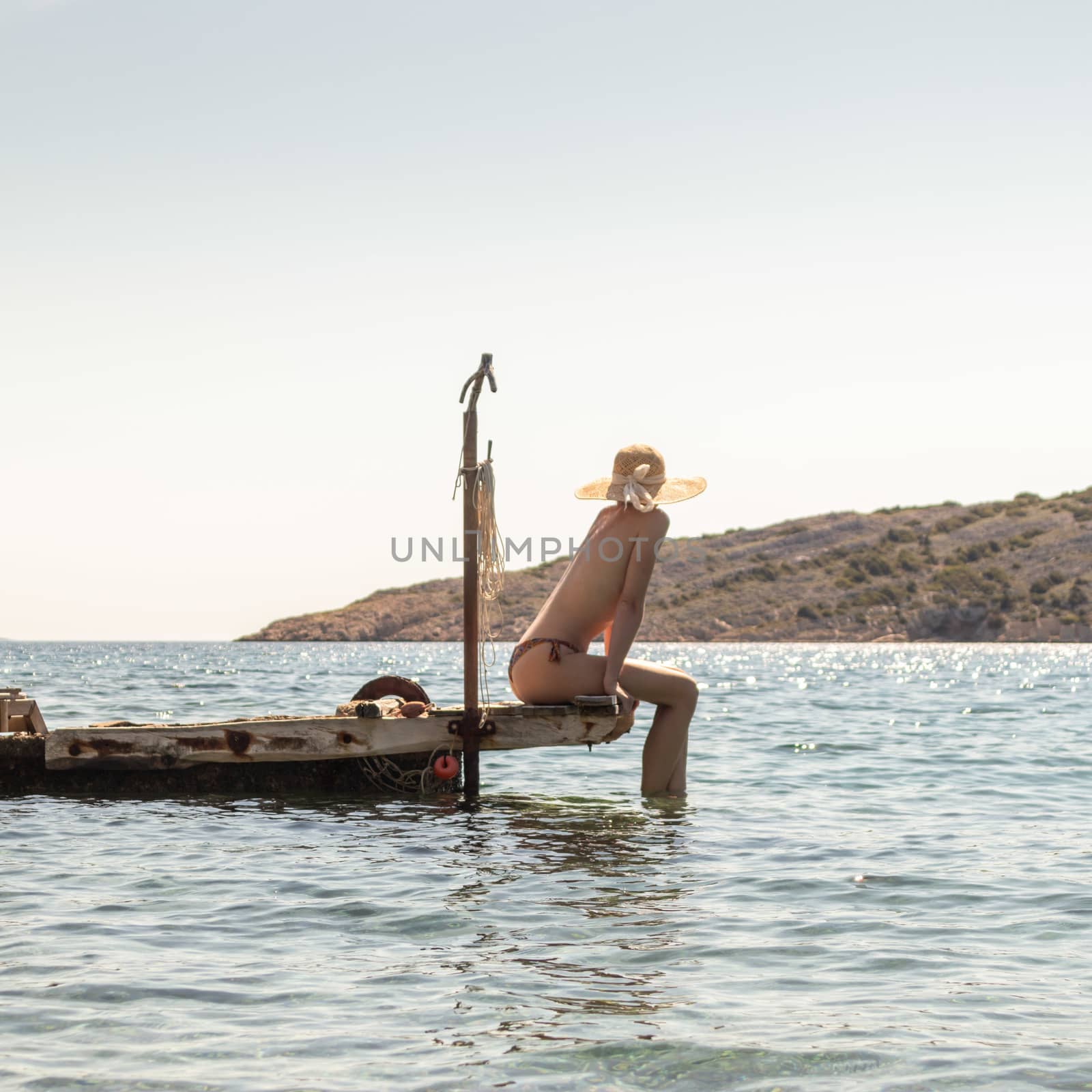 View of unrecognizable woman wearing big summer sun hat tanning topless and relaxing on old wooden pier in remote calm cove of Adriatic sea, Croatia by kasto