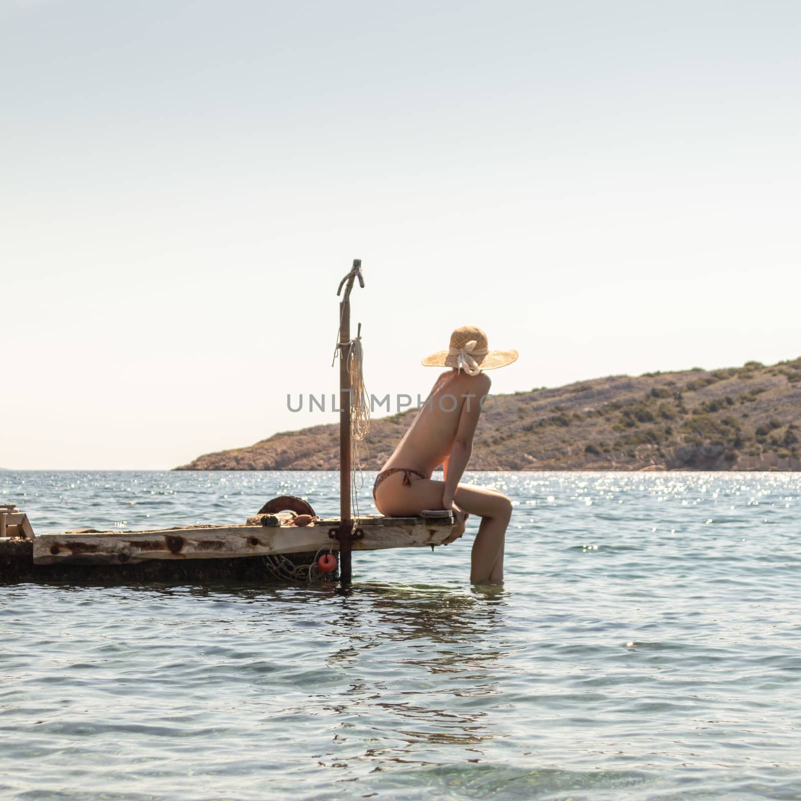 View of unrecognizable woman wearing big summer sun hat tanning topless and relaxing on old wooden pier in remote calm cove of Adriatic sea, Croatia.