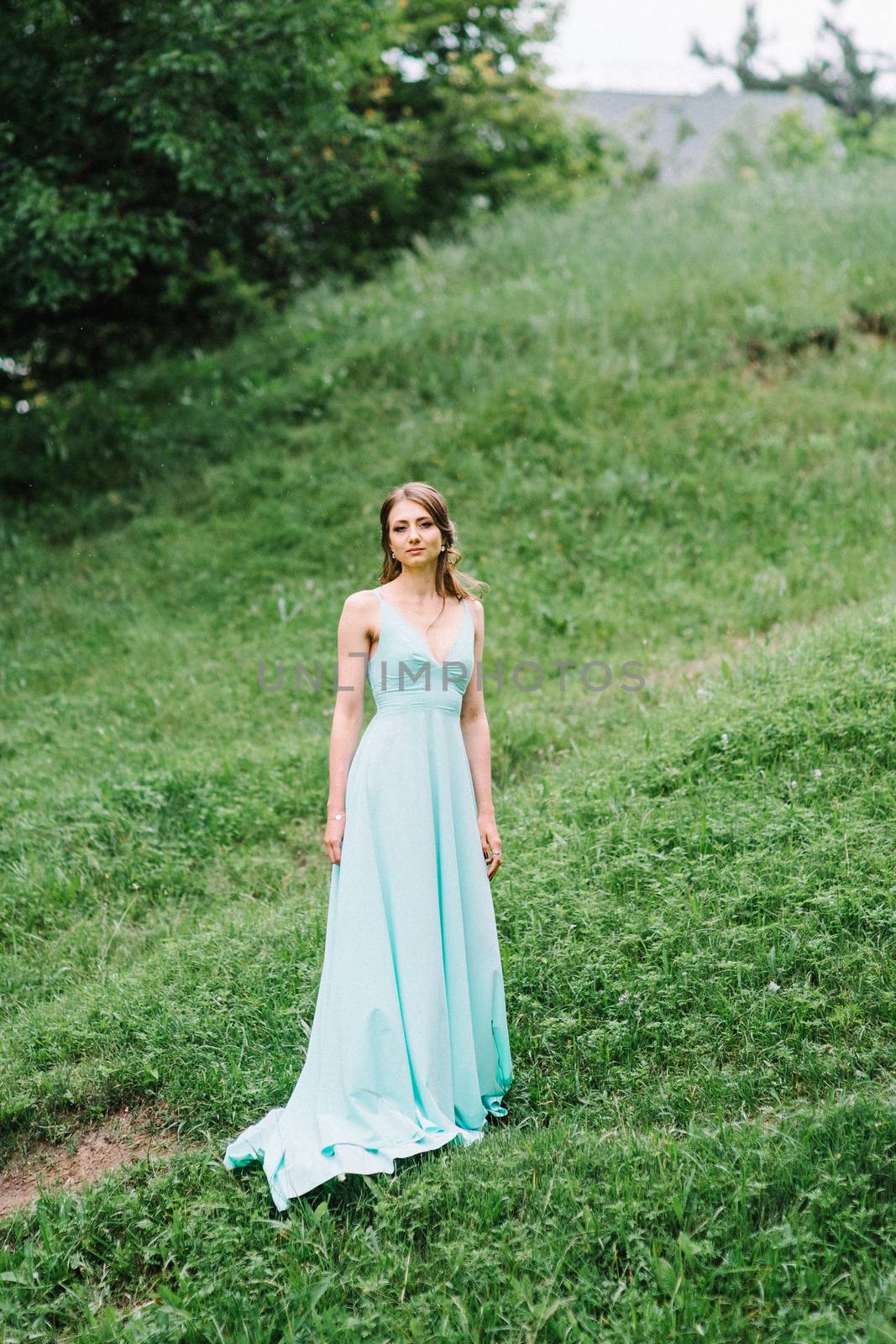 Happy girl in a turquoise long dress in a green park on a background of herbs, trees and rose bushes