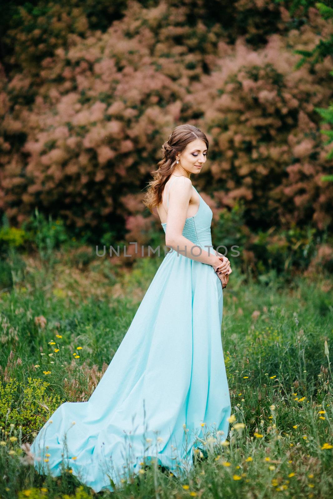 Happy girl in a turquoise long dress in a green park on a background of herbs, trees and rose bushes