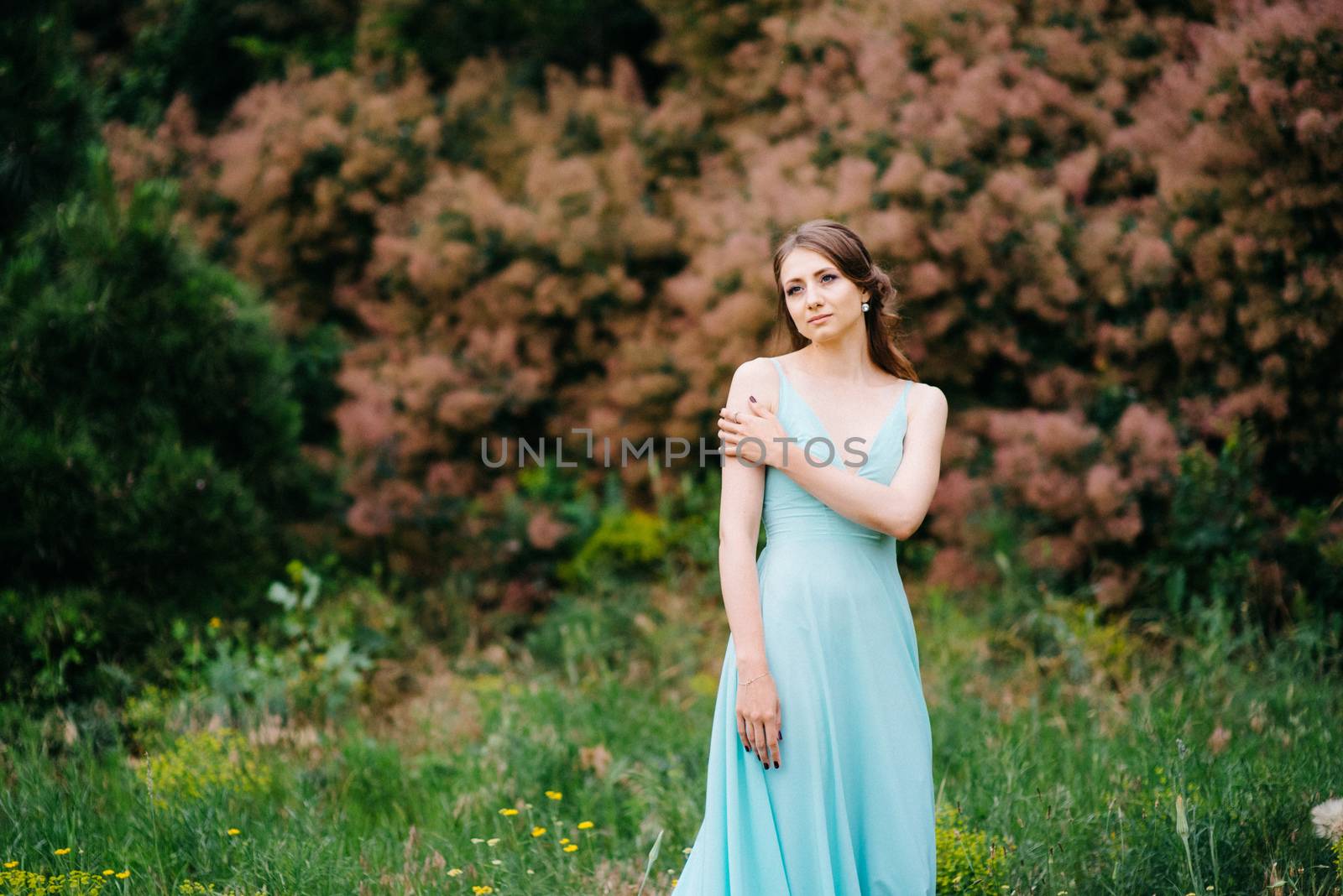 Happy girl in a turquoise long dress in a green park on a background of herbs, trees and rose bushes