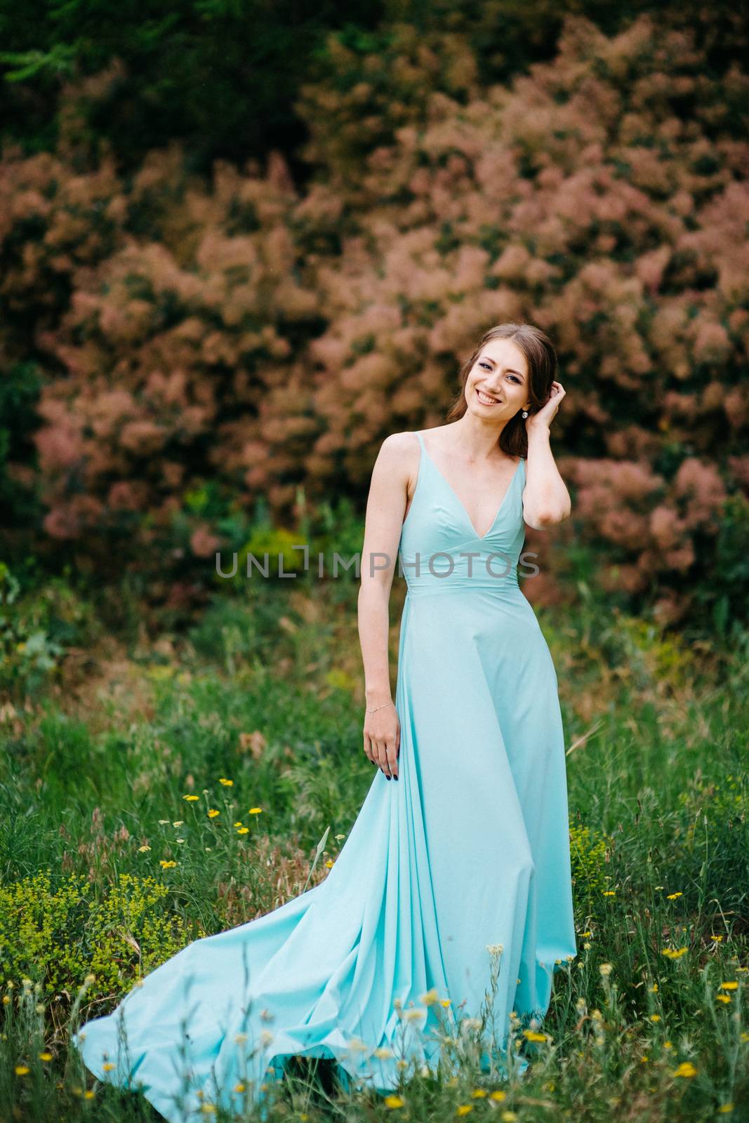 Happy girl in a turquoise long dress in a green park on a background of herbs, trees and rose bushes