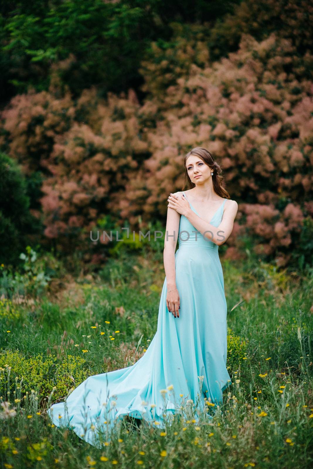 Happy girl in a turquoise long dress in a green park on a background of herbs, trees and rose bushes