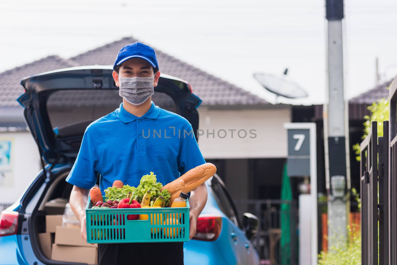 Asian grocery store delivery man wearing blue uniform and face mask protect he delivering fresh food vegetable in plastic box at door front home after coronavirus outbreak, back to new normal concept