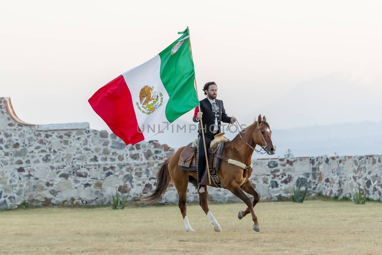 A Very Handsome Mexican Charro Poses In Front Of A Hacienda In The Mexican Countryside While Holding The Mexican Flag by actionsports