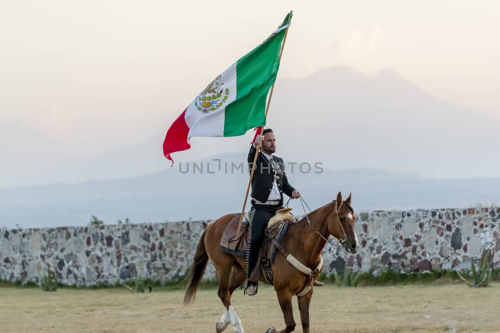 A handsome Mexican Charro poses in front of a hacienda in the Mexican countryside while holding the Mexican Flag