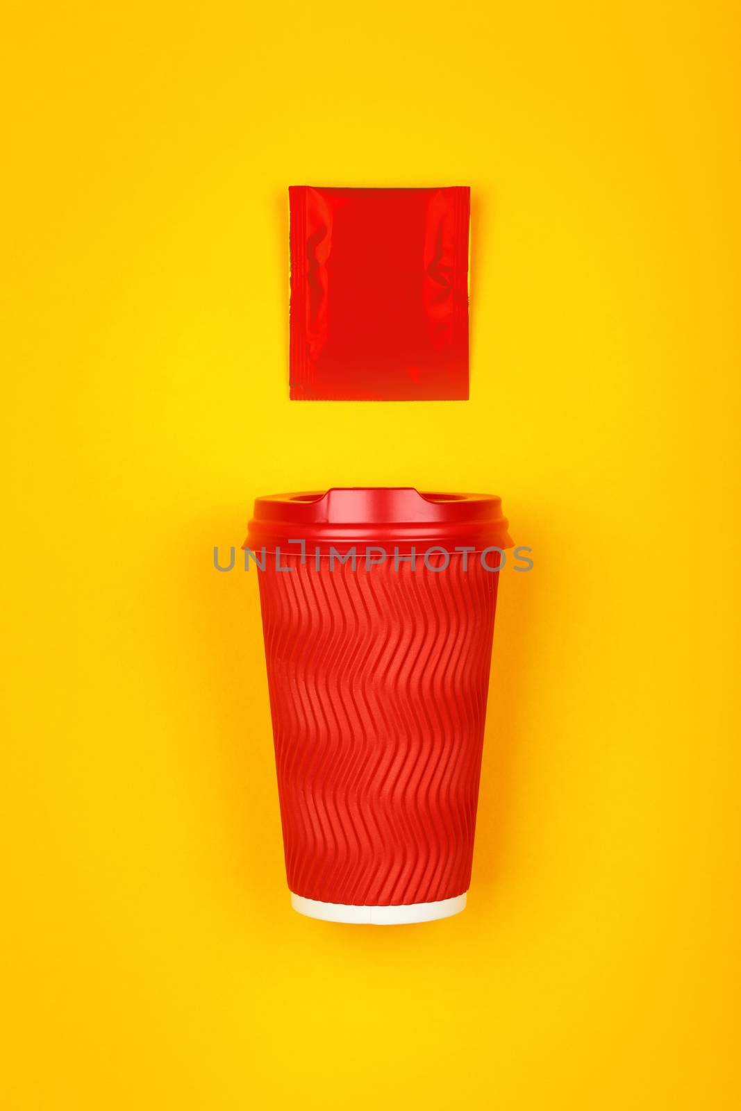 Close up one crimped disposable red paper takeaway cup and instant coffee, sugar or teabag sachet over vivid yellow background, flat lay, elevated top view, directly above
