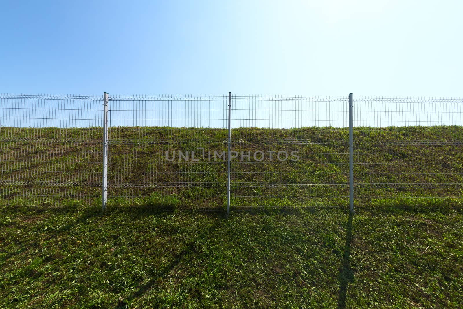 Metal wire industrial grid fence on green grass with blue sky background wide angle horizontal shot.