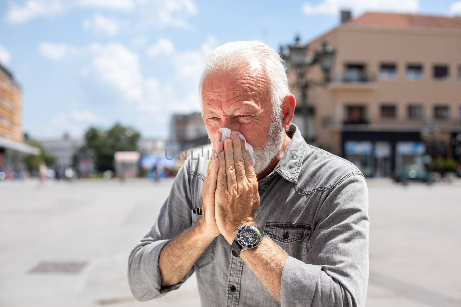 Old man man coughs and sneezes into a handkerchief on street, al by adamr