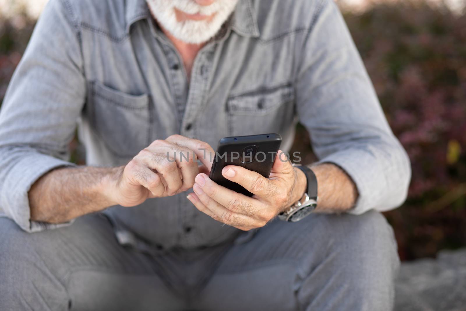 Old man in gray clothing seating on street and typing SMS on smartphone