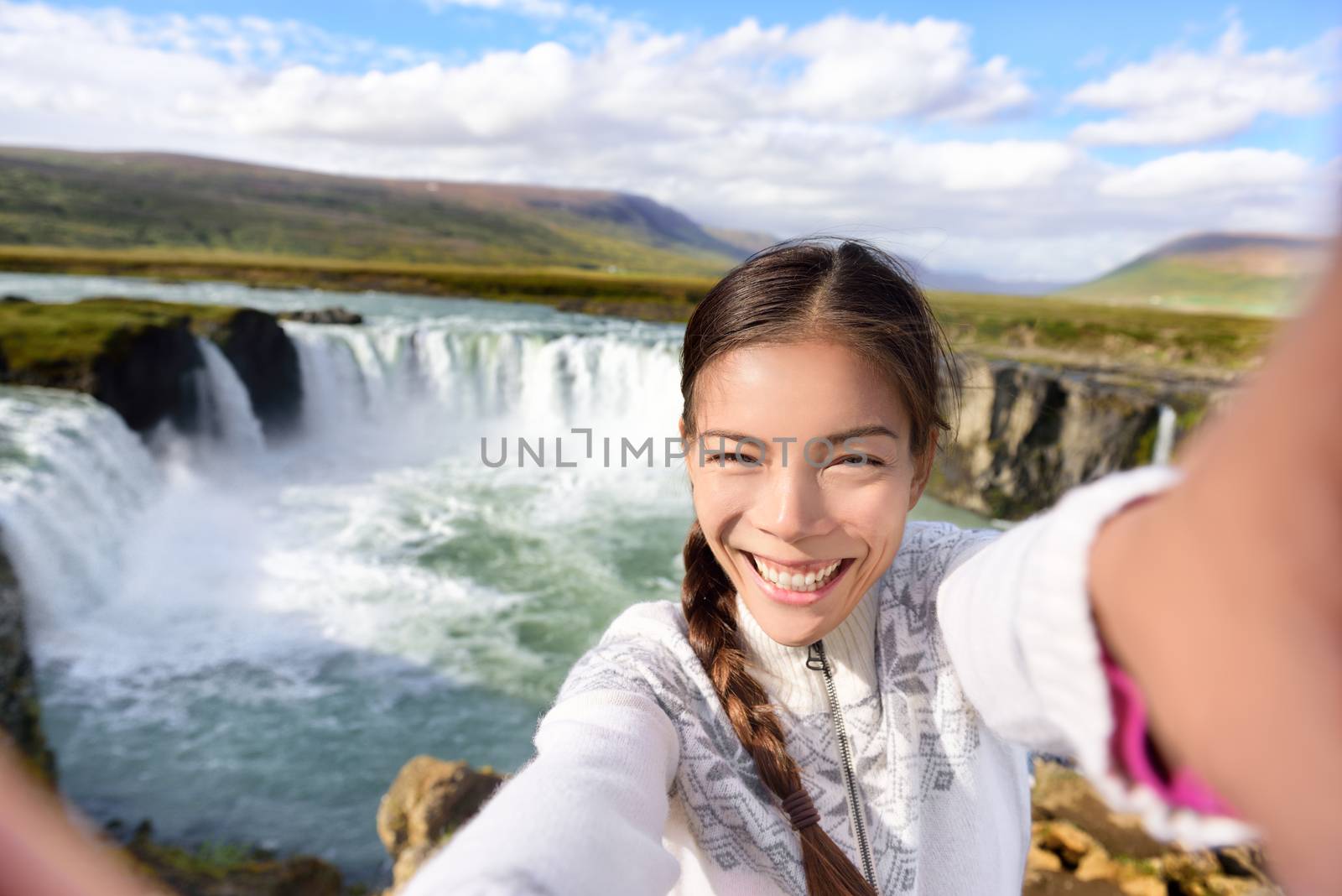 Tourist taking video selfie on travel by Godafoss waterfall on Iceland. Happy young woman tourists enjoying icelandic nature landscape visiting famous tourist destination attraction, Iceland by Maridav