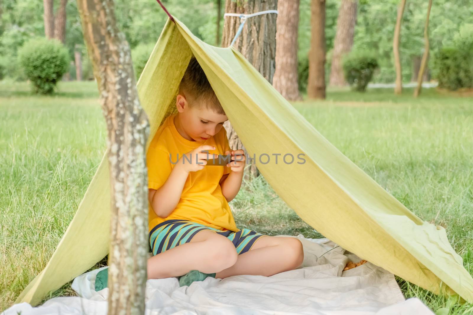 Boy playing video game outdoor in park, backyard, garden. Kid with phone in tent. 6 years old child having fun on nature. E-learning concept. Children and gadgets. Entertainment. Summer holidays by synel