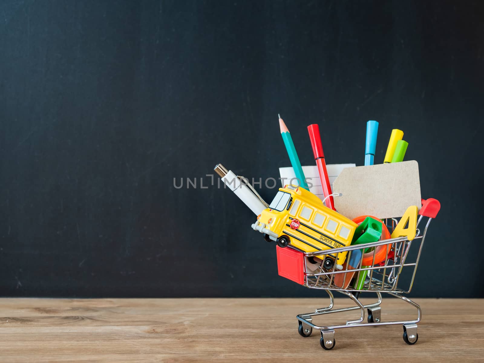 Colorful stationery and school bus model in shopping cart. Back to school concept. Blackboard background with copy space on price tag. School shopping.