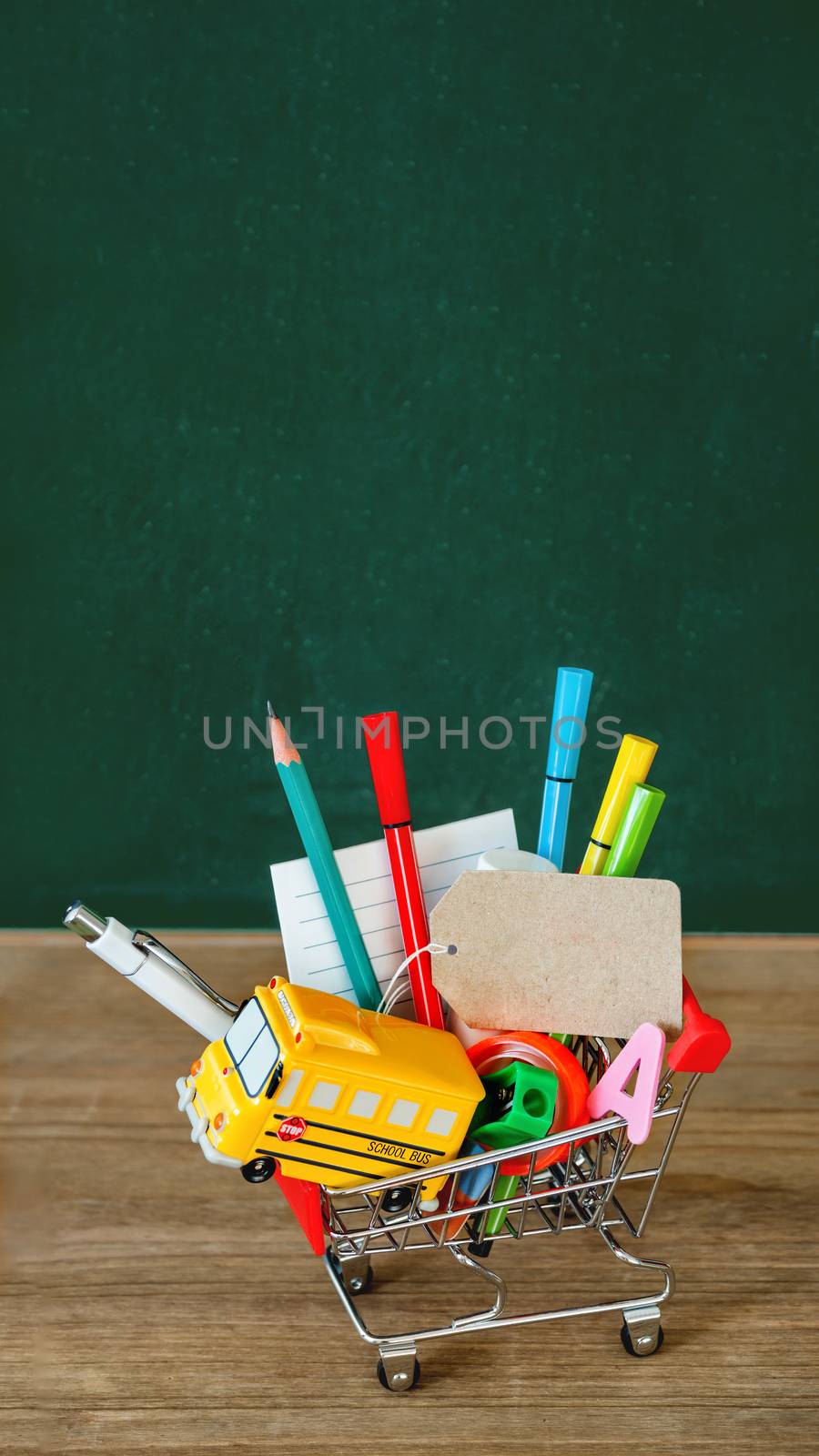 Colorful stationery and school bus model in shopping cart. Back to school concept. Vertical green blackboard background with copy space on tag. 9-16 ratio for mobile applications.