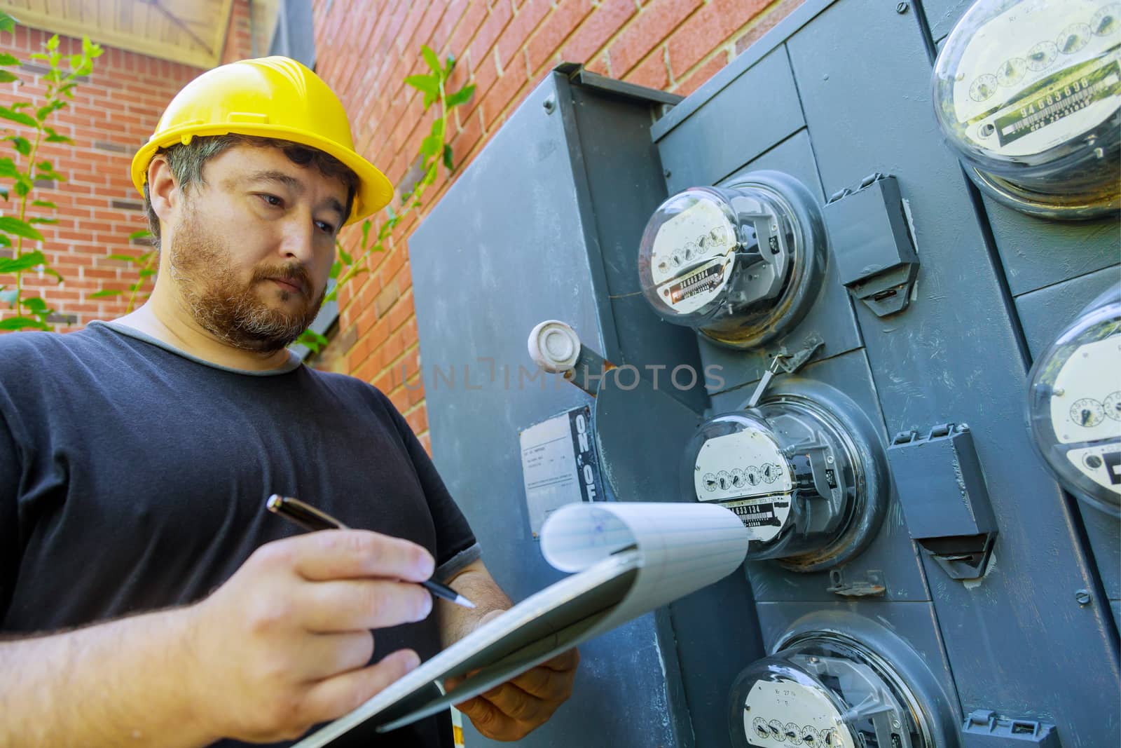 Electrical engineer on during checking in yellow helmet technician examining of doing meter reading on clipboard