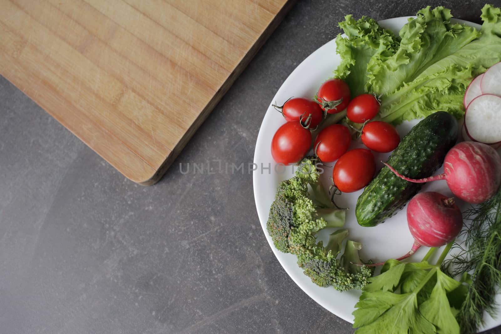 Fresh vegetables in a white plate on a gray table. Tomatoes, cucumber, lettuce, broccoli, radish, cutting board