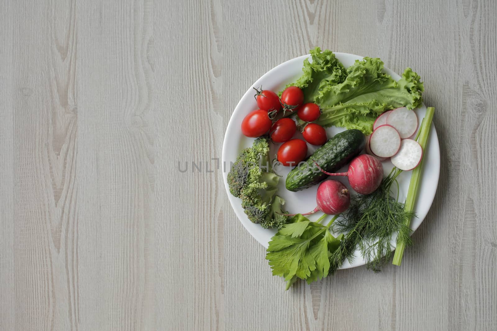 Fresh vegetables in a white plate on a light wooden table. Tomatoes, cucumber, lettuce, broccoli, radish