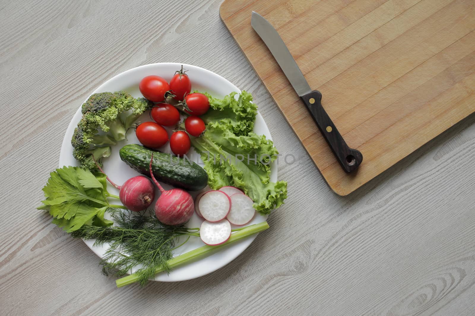 Fresh vegetables in a white plate on a light wooden table. On a plate, tomatoes, cucumber, lettuce, broccoli, radish. Cutting board