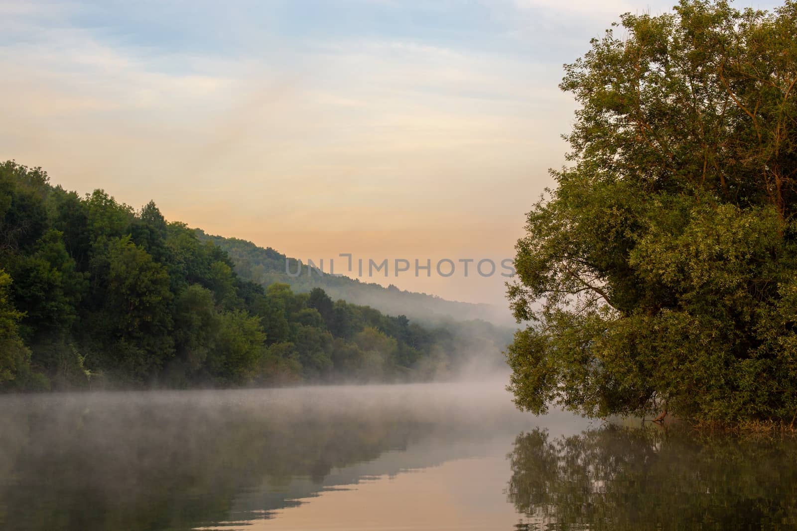 Misty calm small river on a summer morning. Fog floating over the cool water with reflection of the greenery and sky.