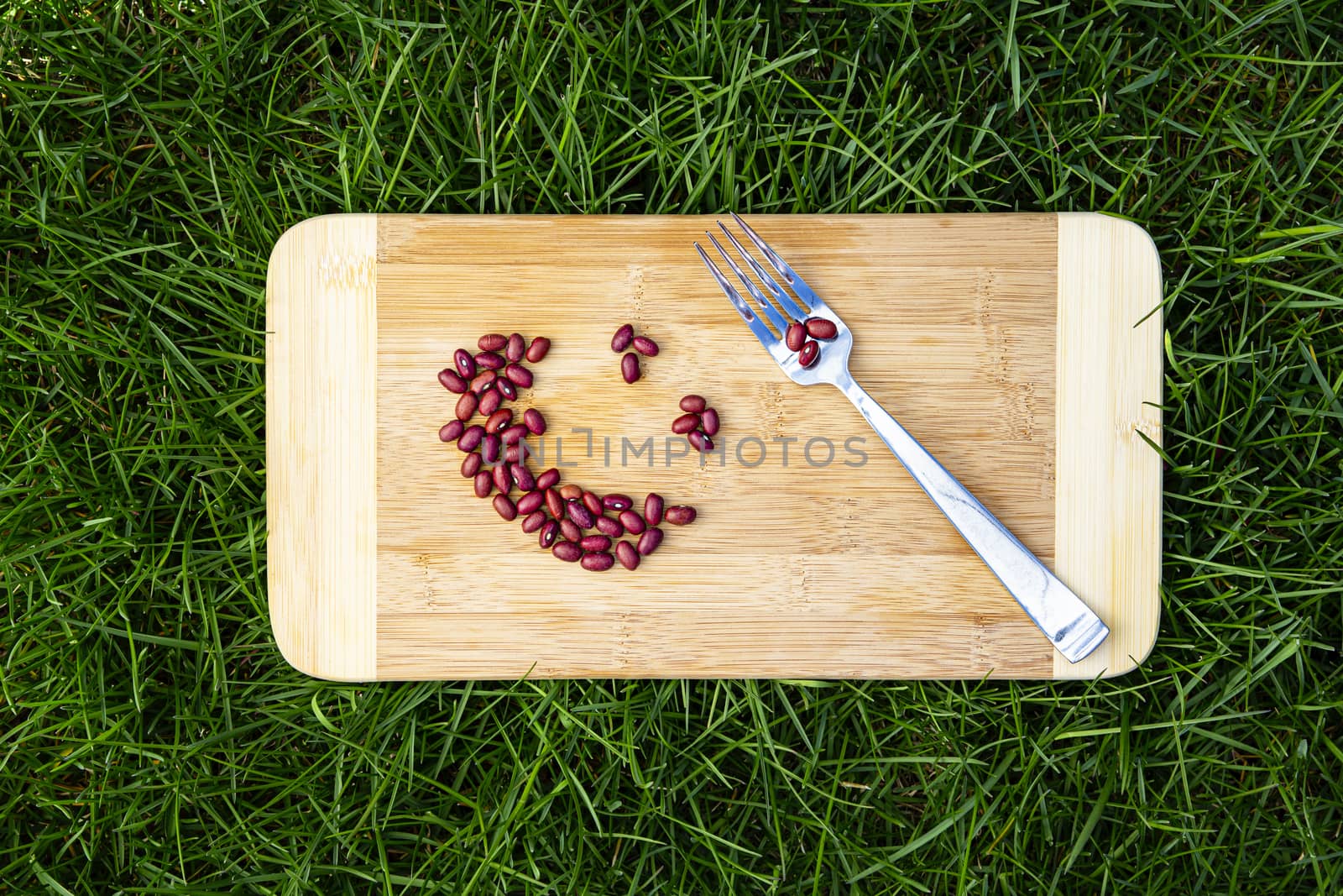 dried red beans, placed in a shape of a smiling face, on top of a wood board, with a metal fork, on top of green grass