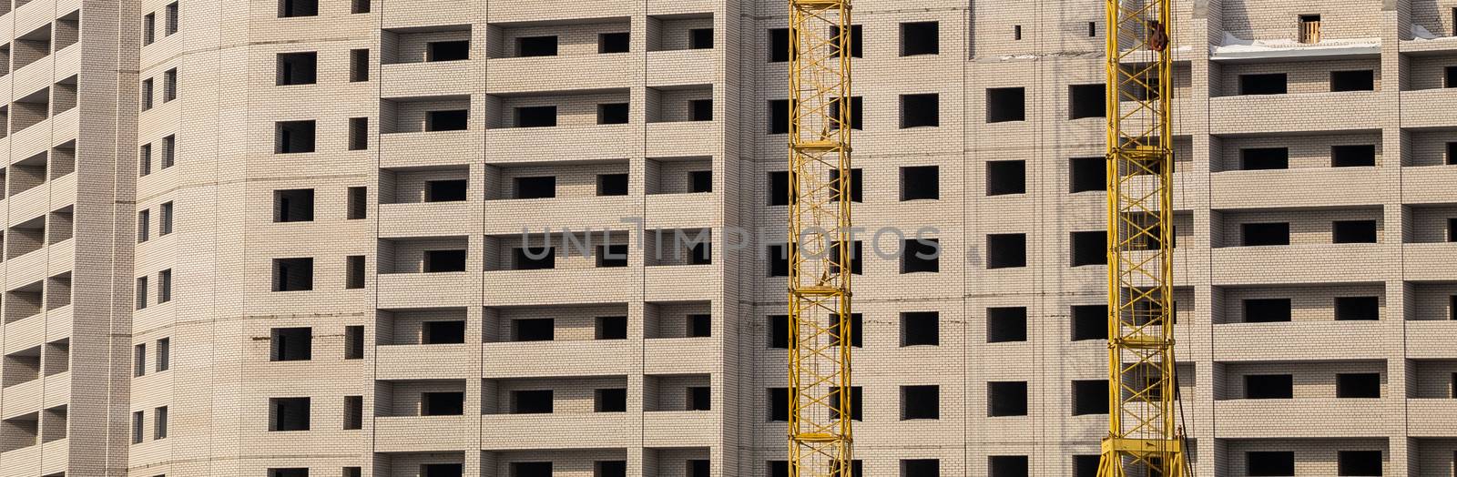 Construction site. Unfinished apartment building made of white bricks. Special yellow industrial construction cranes. Empty windows and balconies.