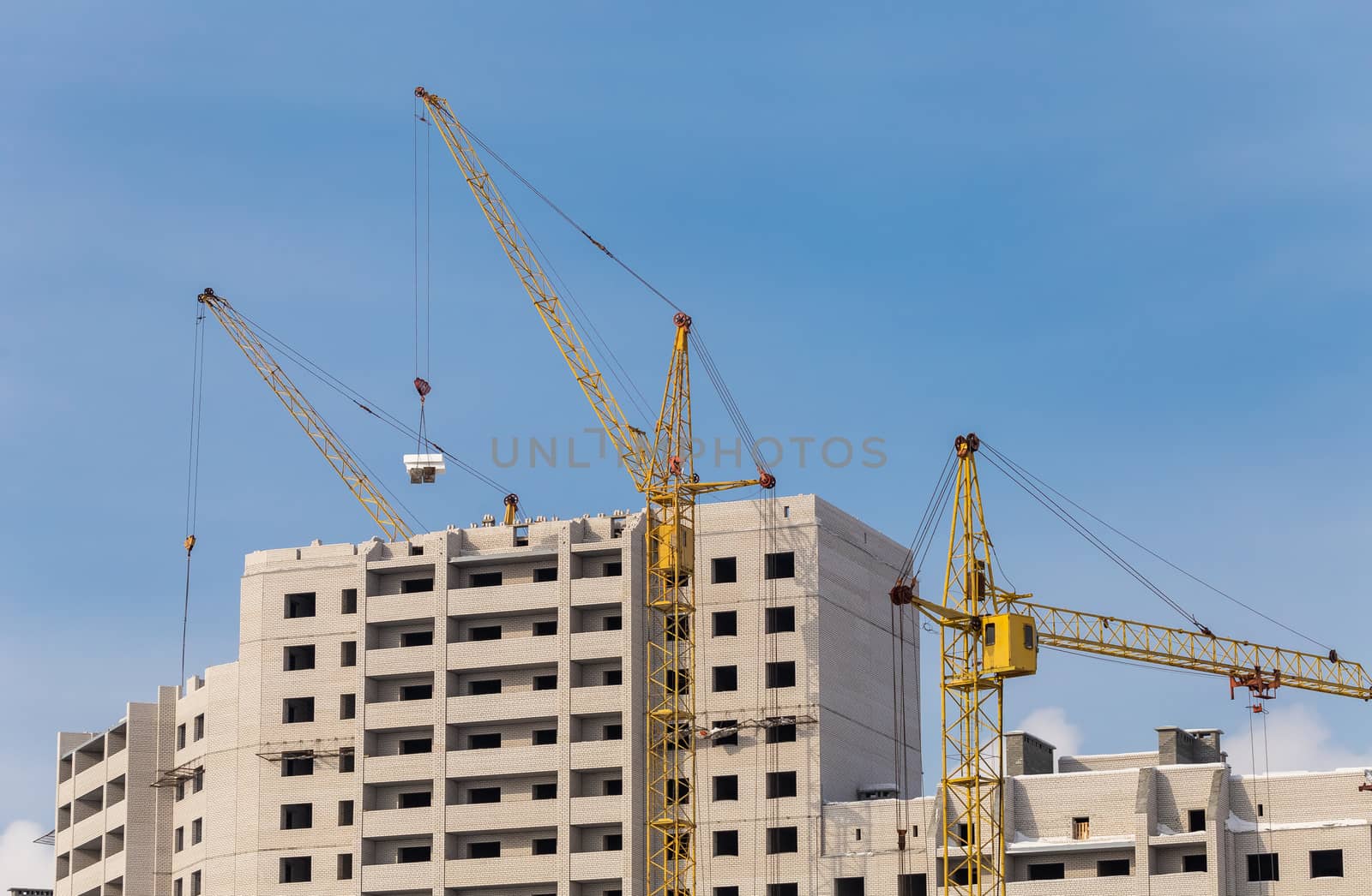 Unfinished apartment buildings. Construction workers laying bricks on top. Special industrial cranes in the middle. Blue sky background