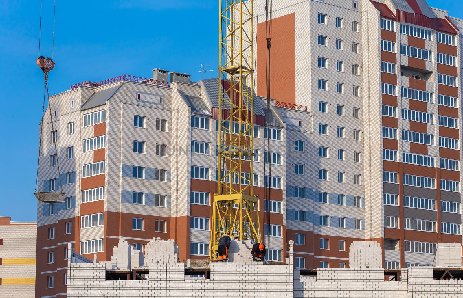 Construction site. Unfinished apartment buildings. Construction workers laying bricks. Special crane in the middle. Blue sky background.