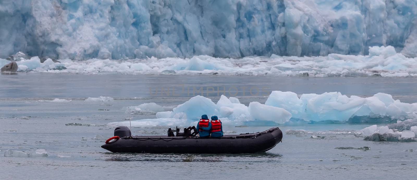 Two people sitting close to each other and wearing life jackets on a boat next to the glacier, icebergs floating around.