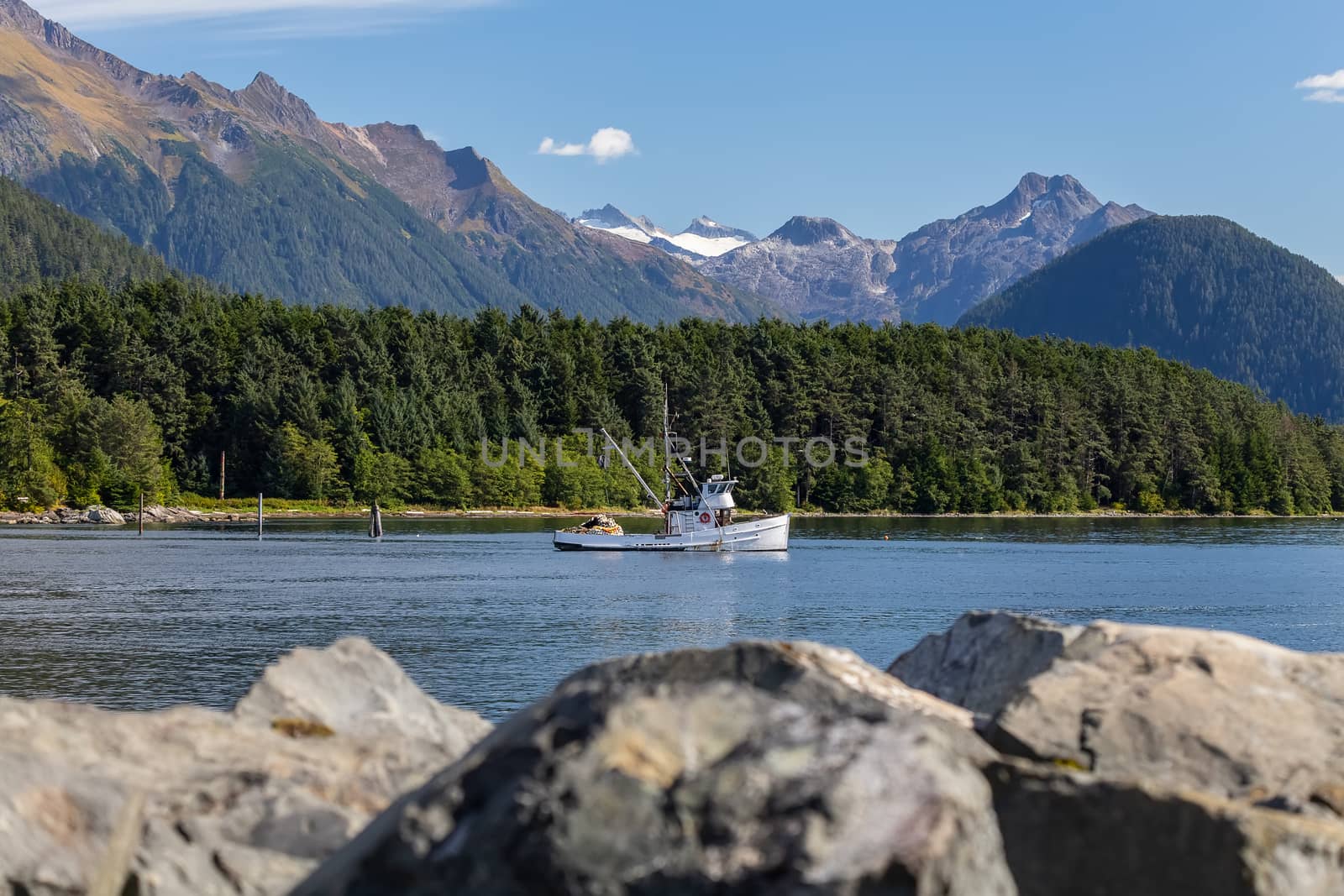 Fishing boat drifting in harbor. Rocks in soft focus in the foreground. Mountains and forest in the background. Sitka, Alaska, USA