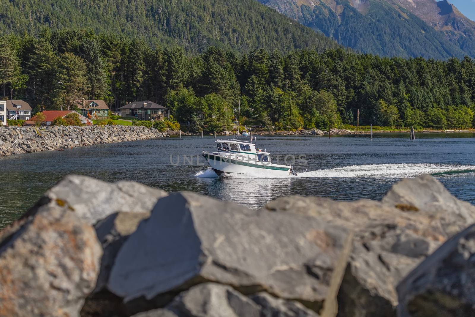 Fishing speedboat sailing fast in harbor. Long water trace behind it. Rocks in soft focus in the foreground. Houses, mountains, and forest in the back