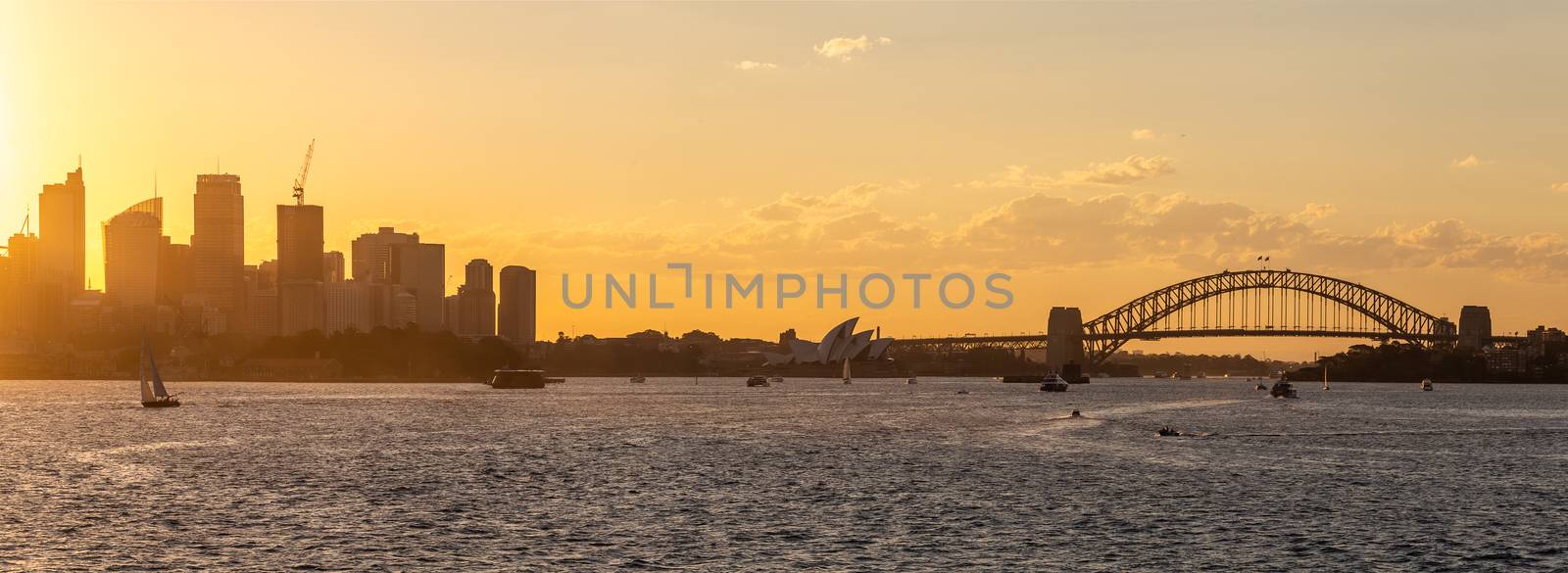Panoramic view of Sydney downtown, harbor bridge and opera house with boats sailing in the bay at sunset. Beautiful orange sky.