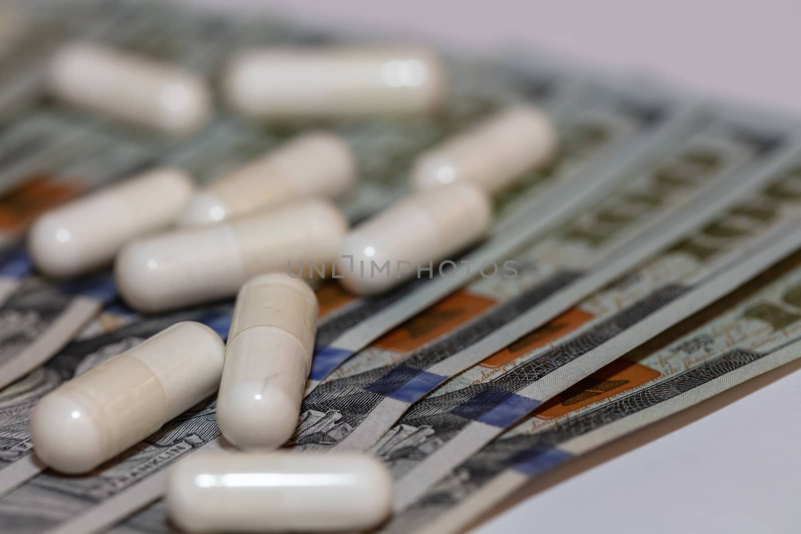 White pills scattered on hundred dollar bills. Majority of the pills and bills are in soft focus. Angled, close-up shot. Healthcare concept