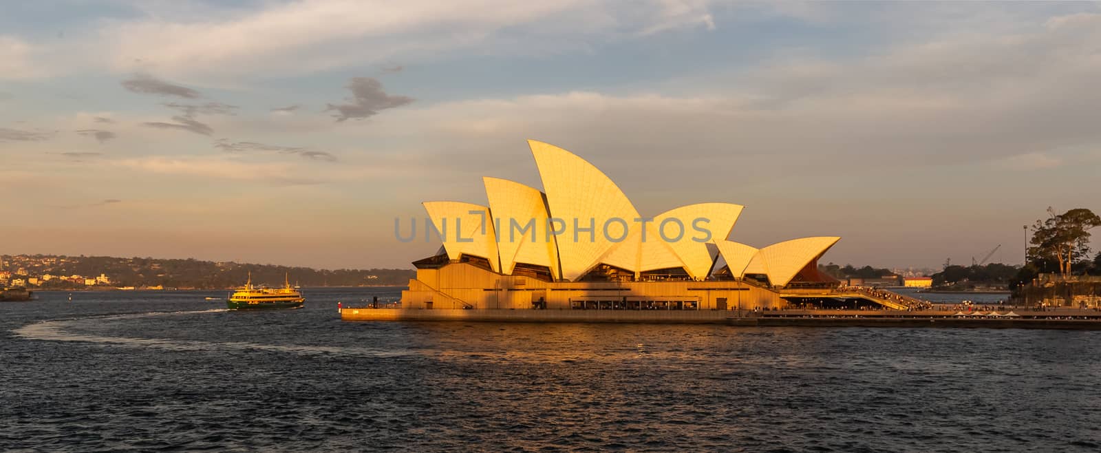 Sydney Harbor, Australia - November 1, 2018: Sydney Opera House at sunset. Boat circling the house in the bay. Beautiful orange-and-yellow colors. Cloudy sky in the background.