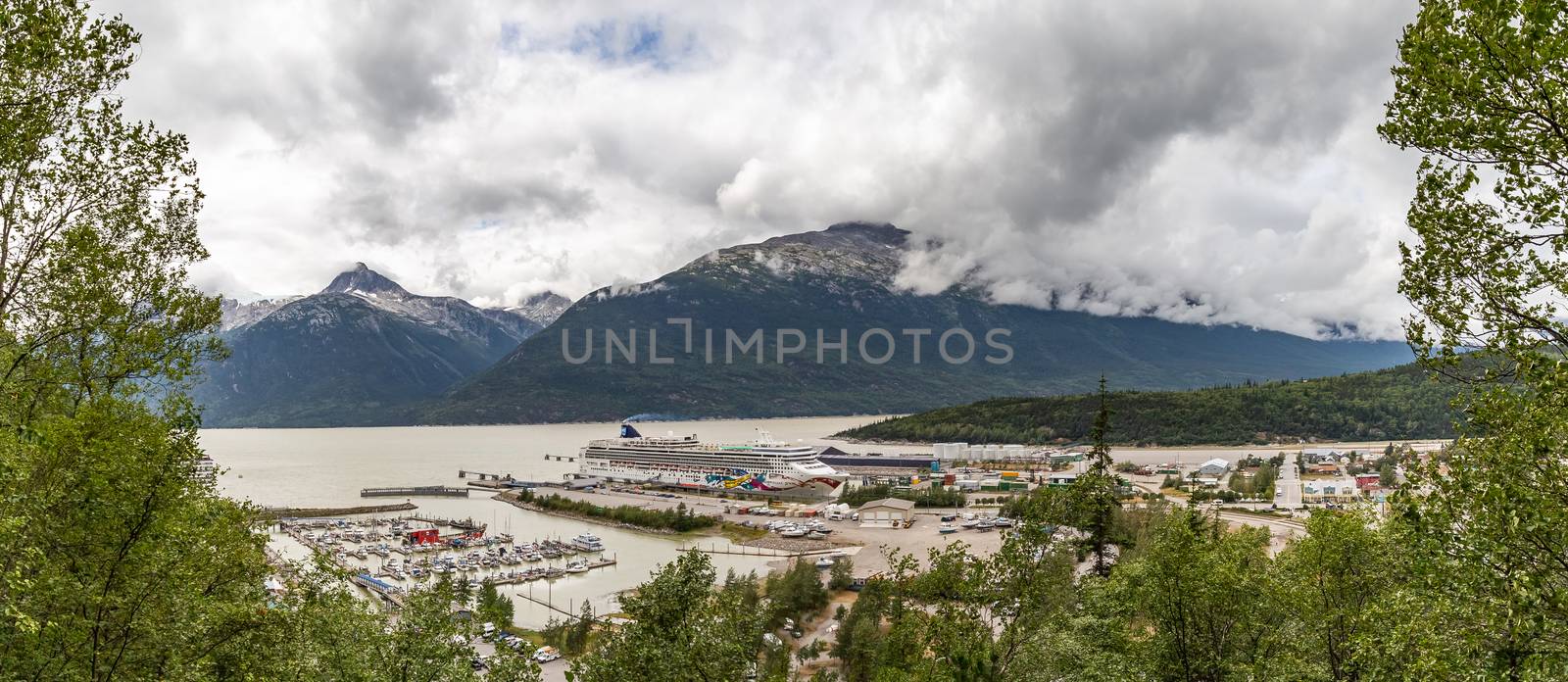 Skagway, Alaska - August 10, 2018: Panoramic view of Skagway port with Norwegian Jewel docked there. Mountains and clouds in the background. Forest in the foreground.