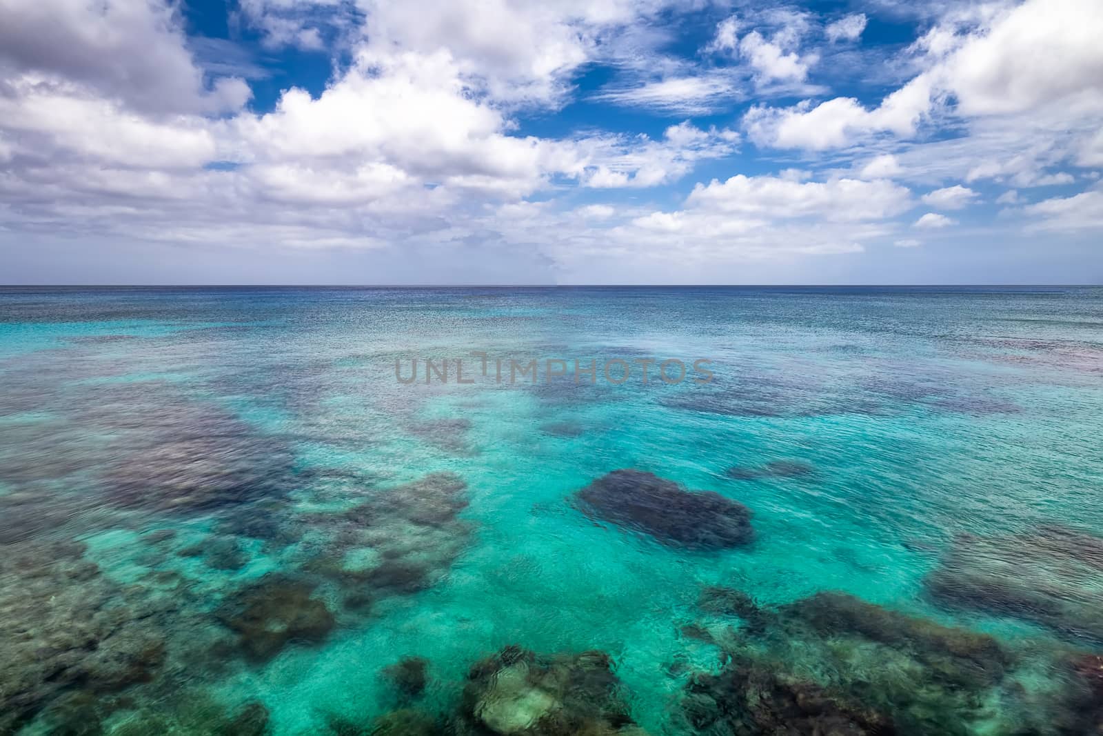Background view of shallow turquoise waters with coral reefs underneath the surface, clear horizon line, blue sky and clouds in the background.