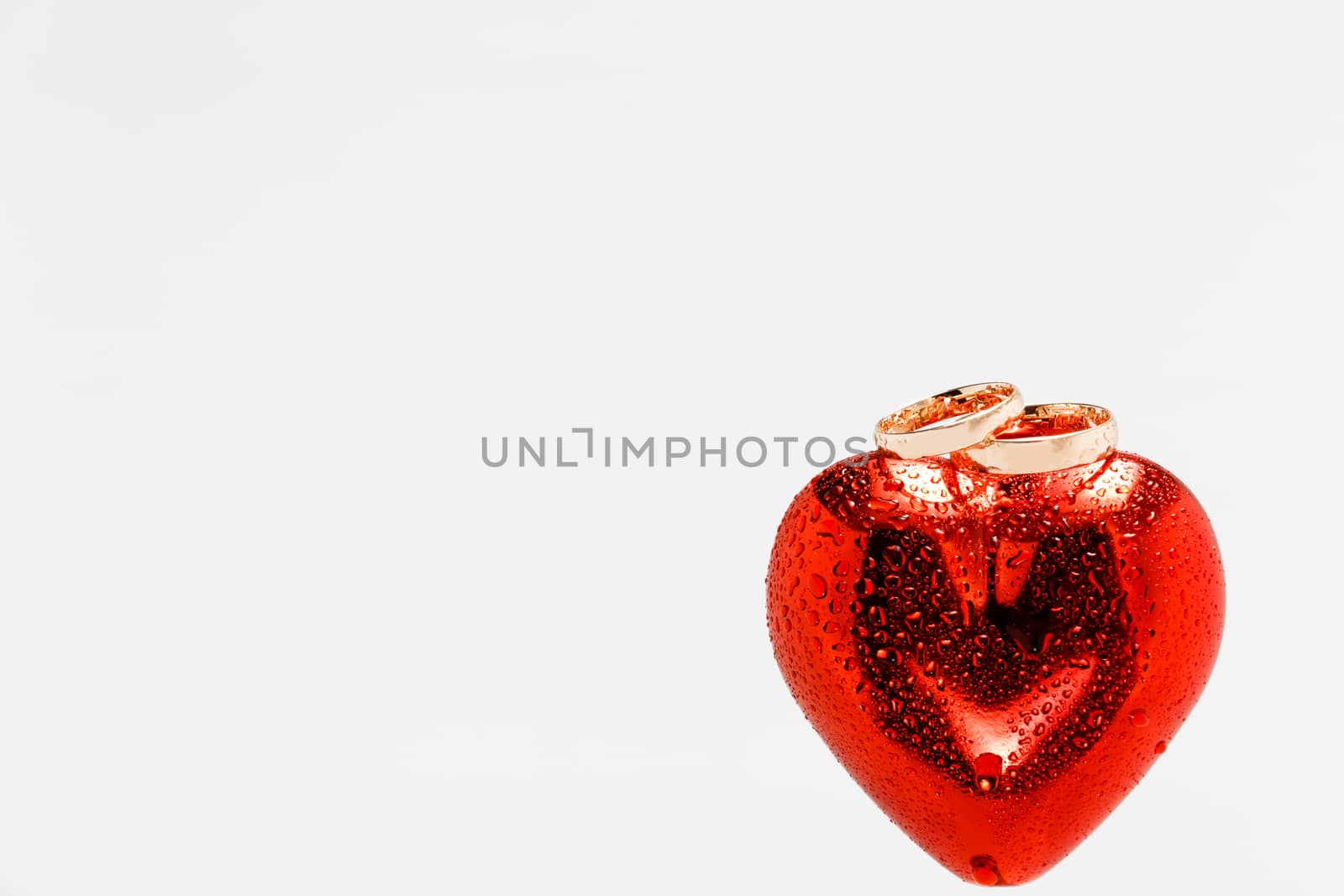 Two gold wedding rings on top of red heart with water drops all over them and the heart. Isolated on white background. Symbol of love and marriage.