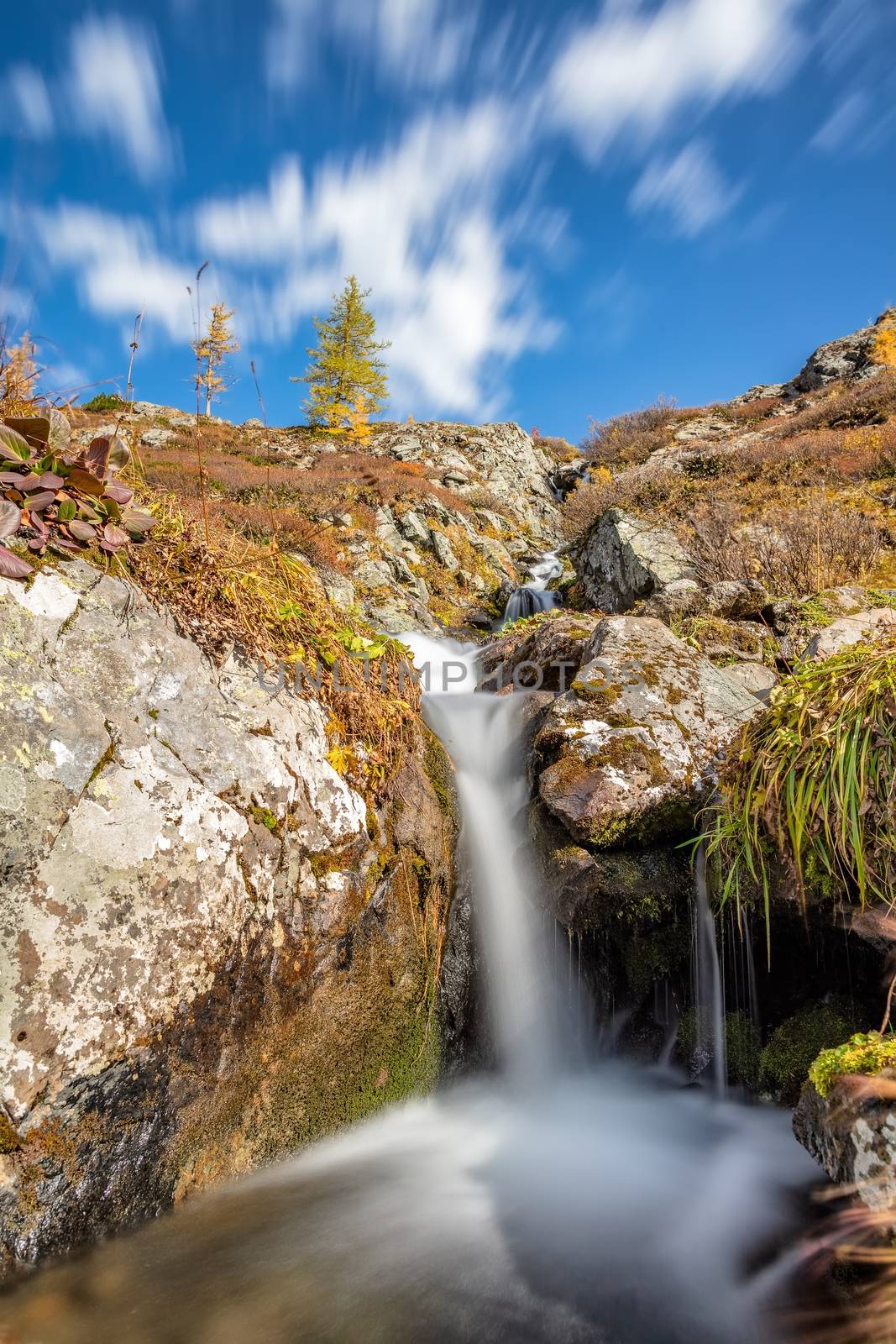 View of a waterfall with smooth water, blue sky and flowing clouds in the background in Altai mountains, Siberia, Russia. Fall 2019. Long exposure