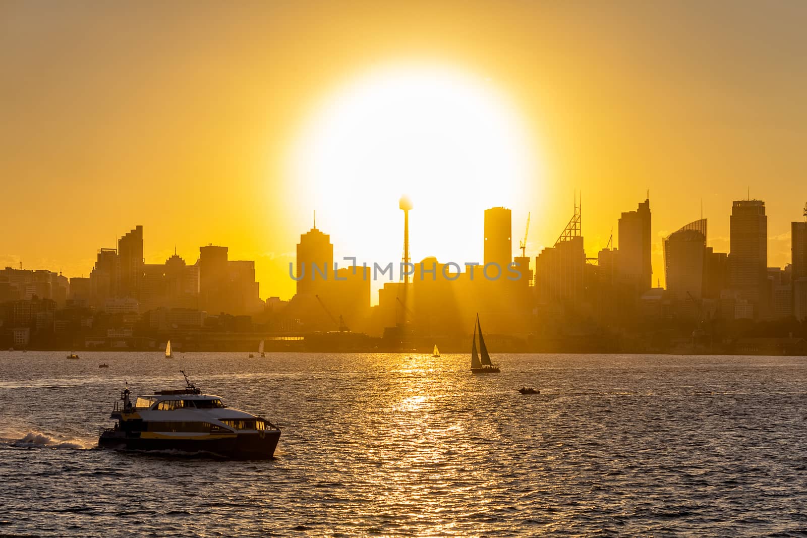 View of downtown Sydney at sunset with Sun disk setting down behind buildings a boat sailing in the foreground