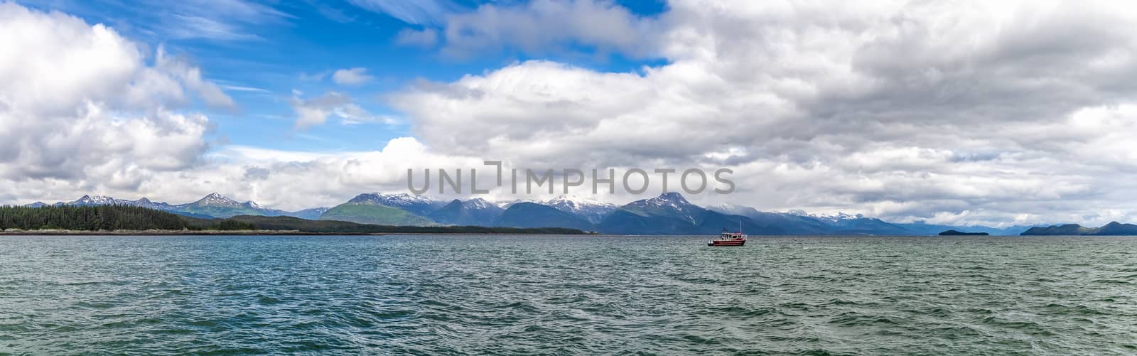 Beautiful panorama with a tiny fishing boat sailing and mountains, clouds and blue sky in the background in Alaska, USA