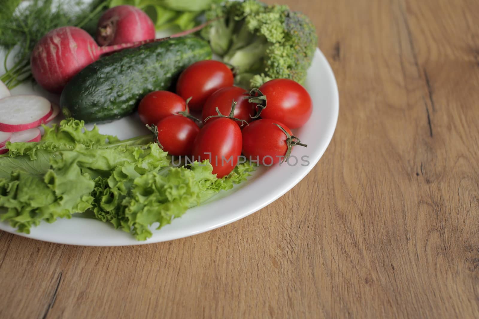 Fresh vegetables in a white plate on a wooden table. Tomatoes, cucumber, lettuce, broccoli, radish