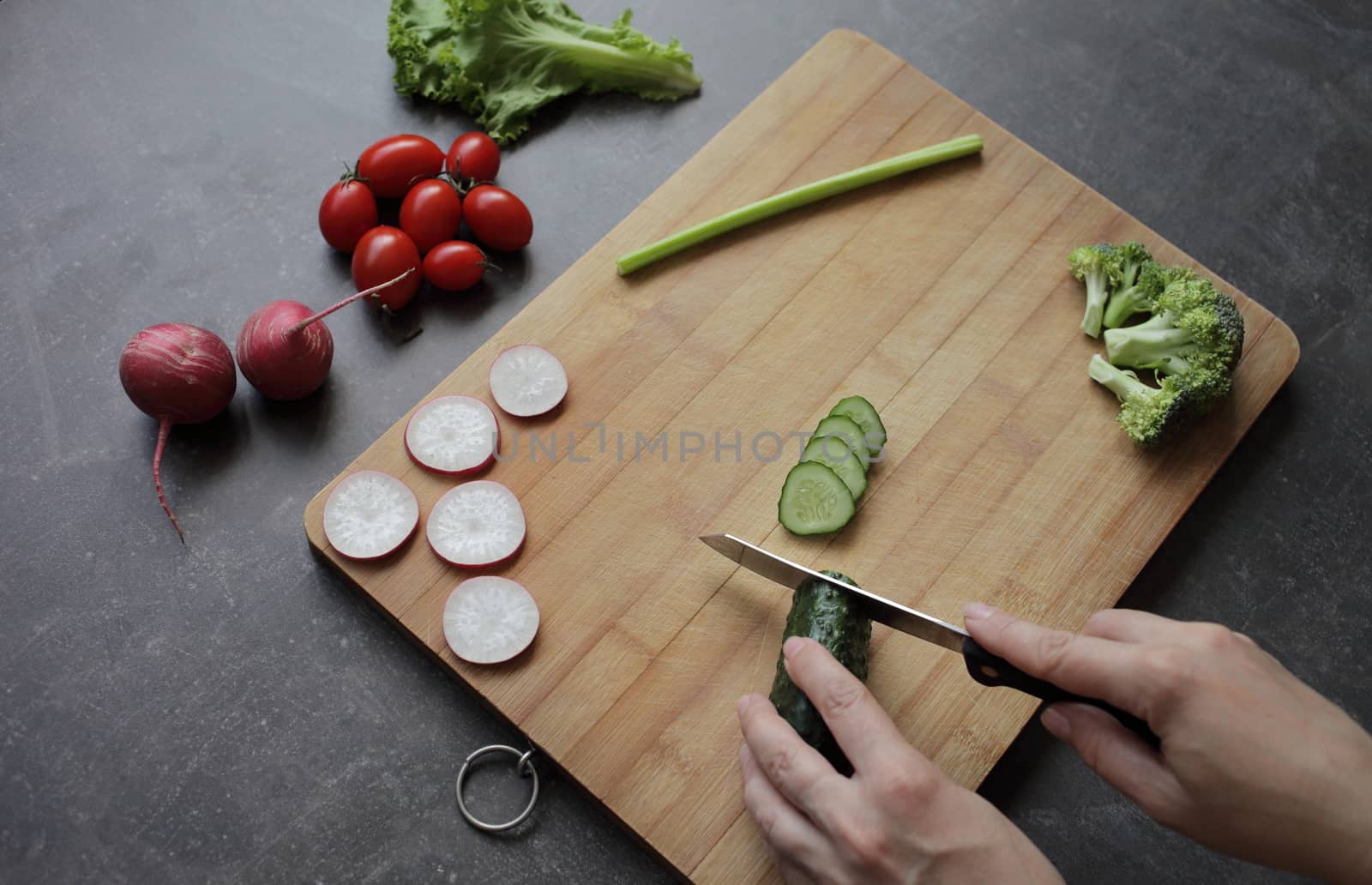 Hands cut cucumber on a cutting board on a gray table. Fresh vegetables, tomatoes, lettuce, broccoli, radishes lie nearby