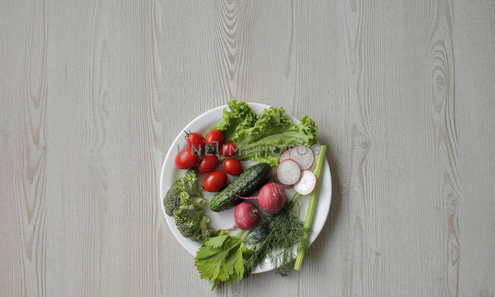 Fresh vegetables in a white plate on a light wooden table. Tomatoes, cucumber, lettuce, broccoli, radish