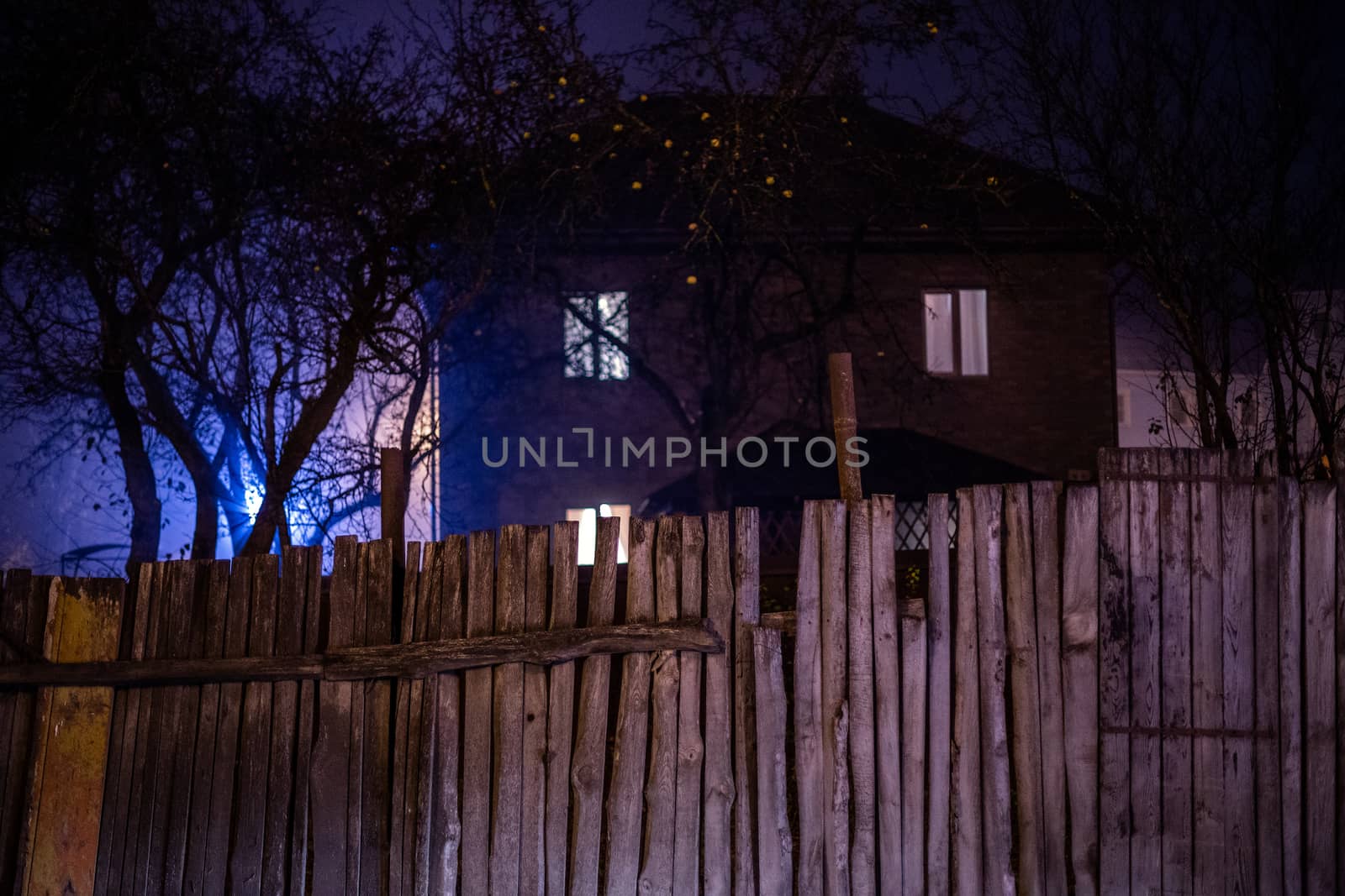 Dark foggy night view on wooden fence and spooky house surrounded by black dry apple trees with selective focus and shallow depth of field.